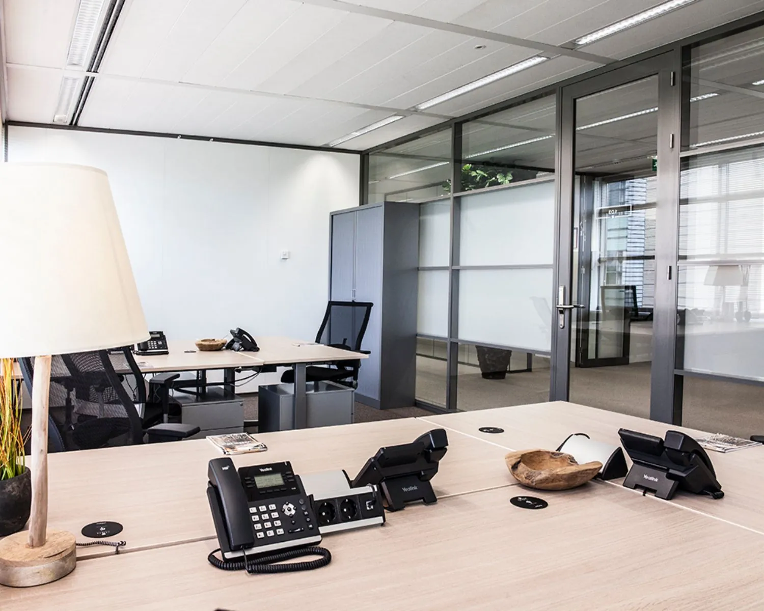 Modern en licht ingericht kantoor met lege bureaus, telefoons en glazen scheidingswanden.