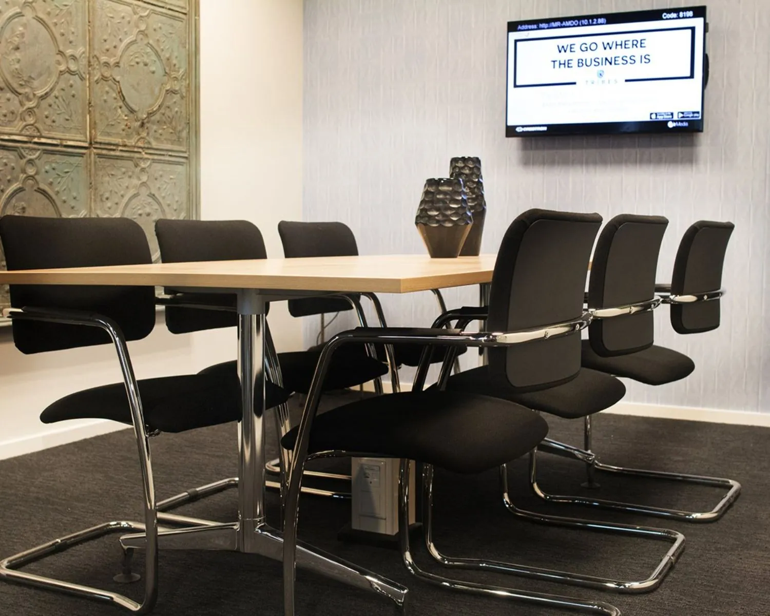 Vergaderruimte met een houten tafel, acht zwarte stoelen, decoratieve vazen en een scherm aan de muur met de tekst “We go where the business is”.