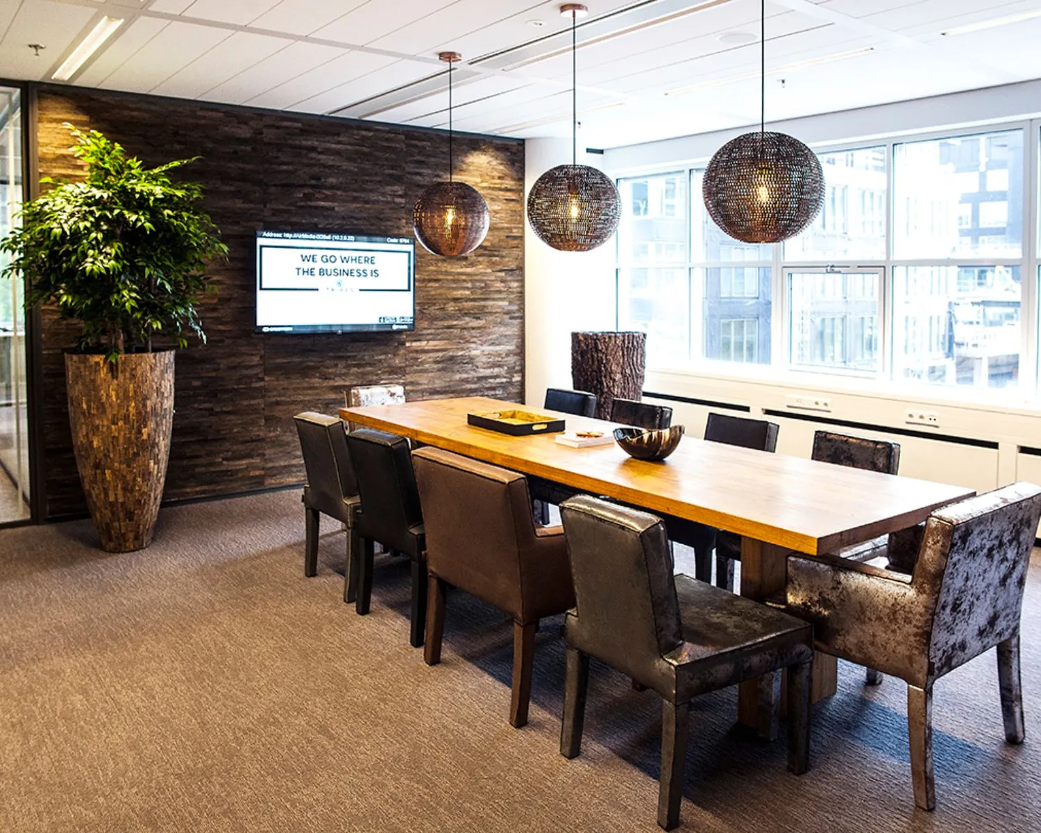 Moderne vergaderruimte met lange houten tafel, diverse stoelen, hanglampen en een wand met tv-scherm.