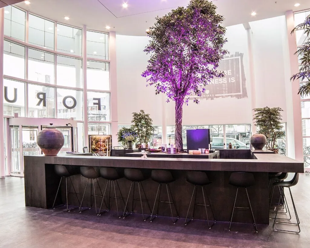 Moderne lounge met hoge tafels, zwarte barkrukken en een verlicht kunstboom in het midden van een lichte ruimte met grote ramen.