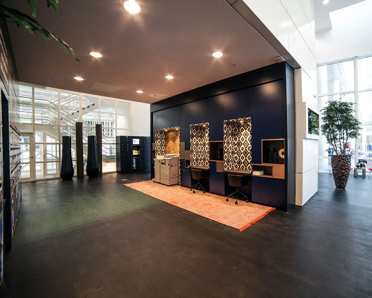 Moderne gemeenschappelijke werkruimte met bureaus, een printer, kluisjes en grote planten in een open en lichte omgeving.