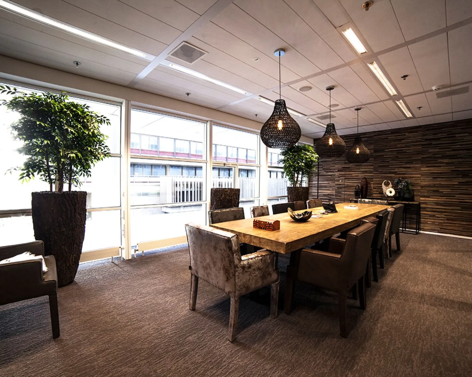 Vergaderruimte met lange houten tafel, moderne stoelen, hanglampen en grote ramen met uitzicht naar buiten.