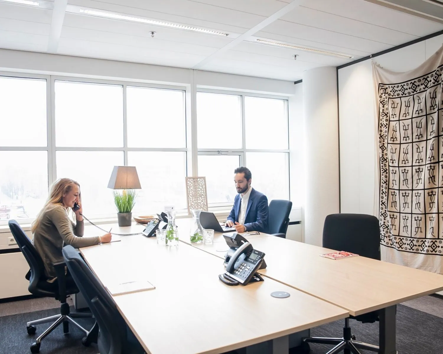 Twee mensen werken in een modern kantoor met grote ramen en een decoratief wandkleed.