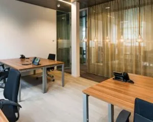 Moderne kantoorruimte met houten bureaus, zwarte bureaustoelen, telefoontoestellen en hanglampen voor een glazen wand met gordijnen.