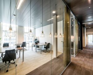 Modern glazen kantoor met meerdere bureaus, bureaustoelen en hanglampen, verlicht door natuurlijk daglicht.