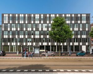 Modern kantoorgebouw met een strakke gevel en verticale ramen, gelegen aan een straat met tramrails, bushalte en voorbijgangers.