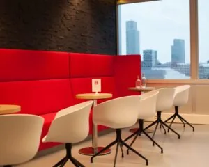 Moderne zithoek met rode wandbank, witte stoelen en uitzicht op een stedelijke skyline.