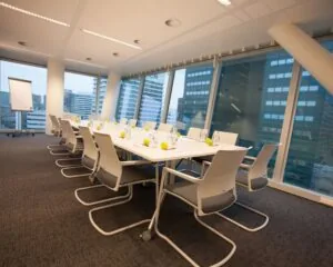 Moderne vergaderruimte met een lange witte tafel, witte stoelen, en uitzicht op hoge kantoorgebouwen.