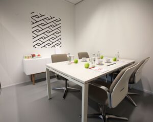 Vergaderruimte met een witte tafel, zes stoelen, groene appels, kopjes en notitieblokken, en een modern kunstwerk aan de muur.