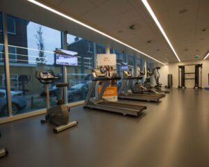 Moderne fitnessruimte met cardioapparatuur, televisieschermen en grote ramen met uitzicht op de straat.