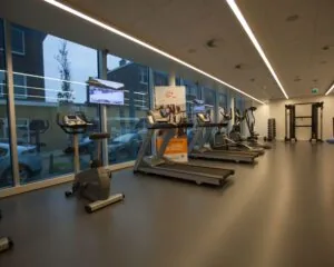 Moderne fitnessruimte met cardioapparatuur, televisieschermen en grote ramen met uitzicht op de straat.