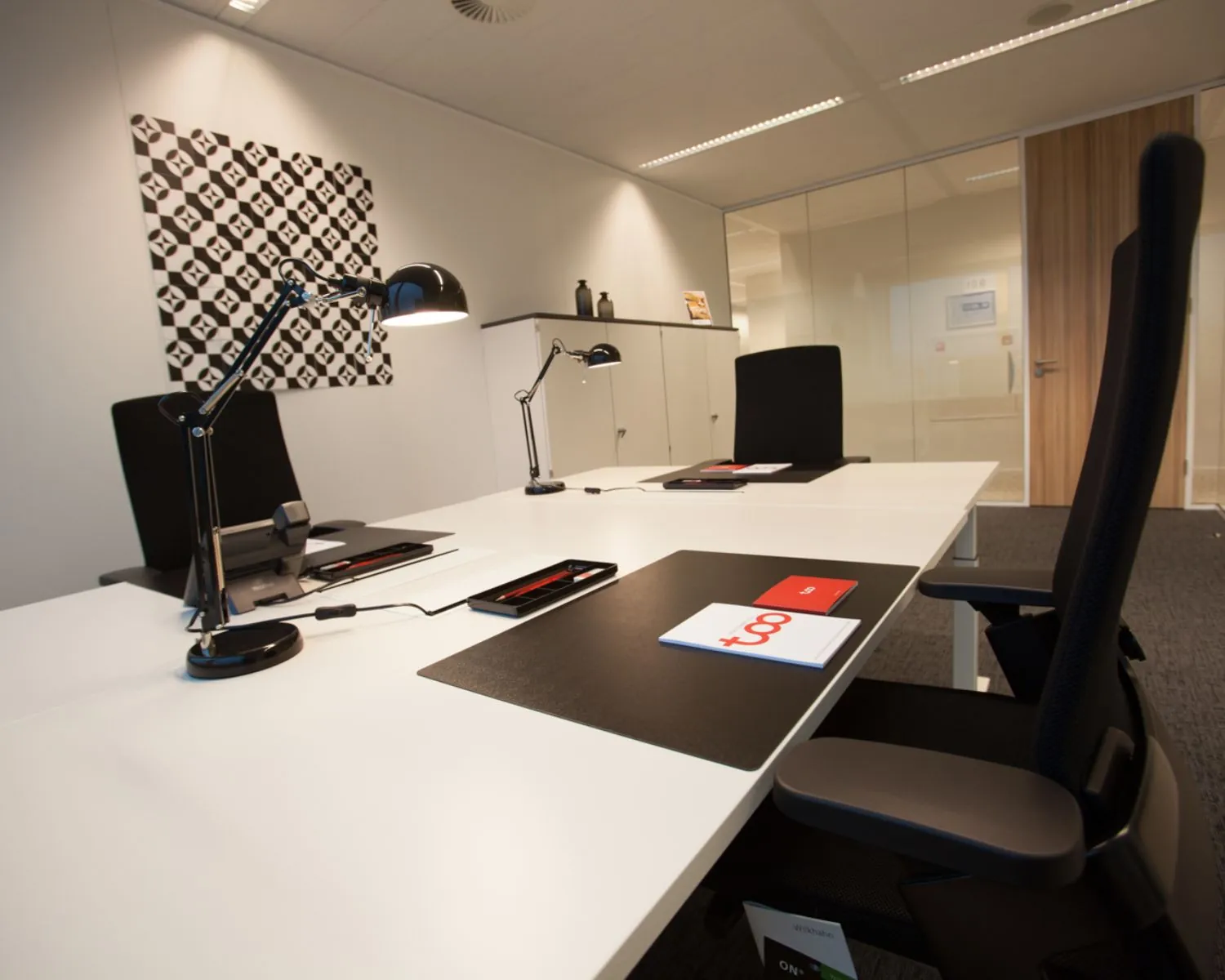 Moderne kantoorruimte met vier werkplekken, bureaulampen en zwarte stoelen, tegen een witte muur met grafische kunst.