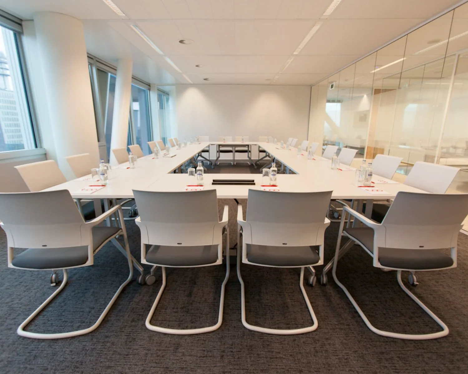 Moderne vergaderruimte met een U-vormige opstelling van witte tafels en stoelen in een kantooromgeving.