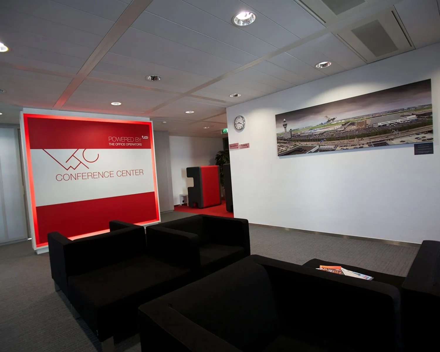Moderne wachtruimte van een conferentiecentrum met zwarte zitbanken, een rode ontvangstwand en een grote foto van luchthaven Schiphol aan de muur.