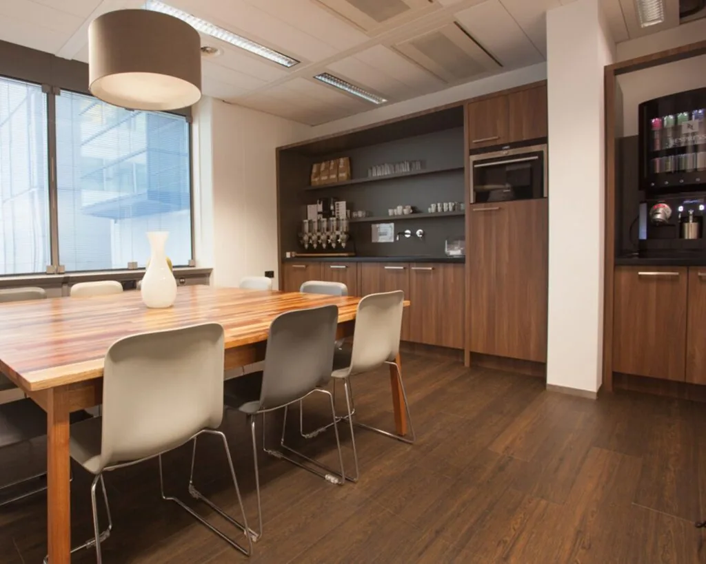 Moderne kantoorkeuken met houten vloer, lange vergadertafel, grijze stoelen en ingebouwde koffiemachines.