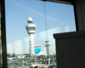 Uitzicht op de verkeerstoren van Schiphol Airport door een raam met op de voorgrond een parkeerplaats.