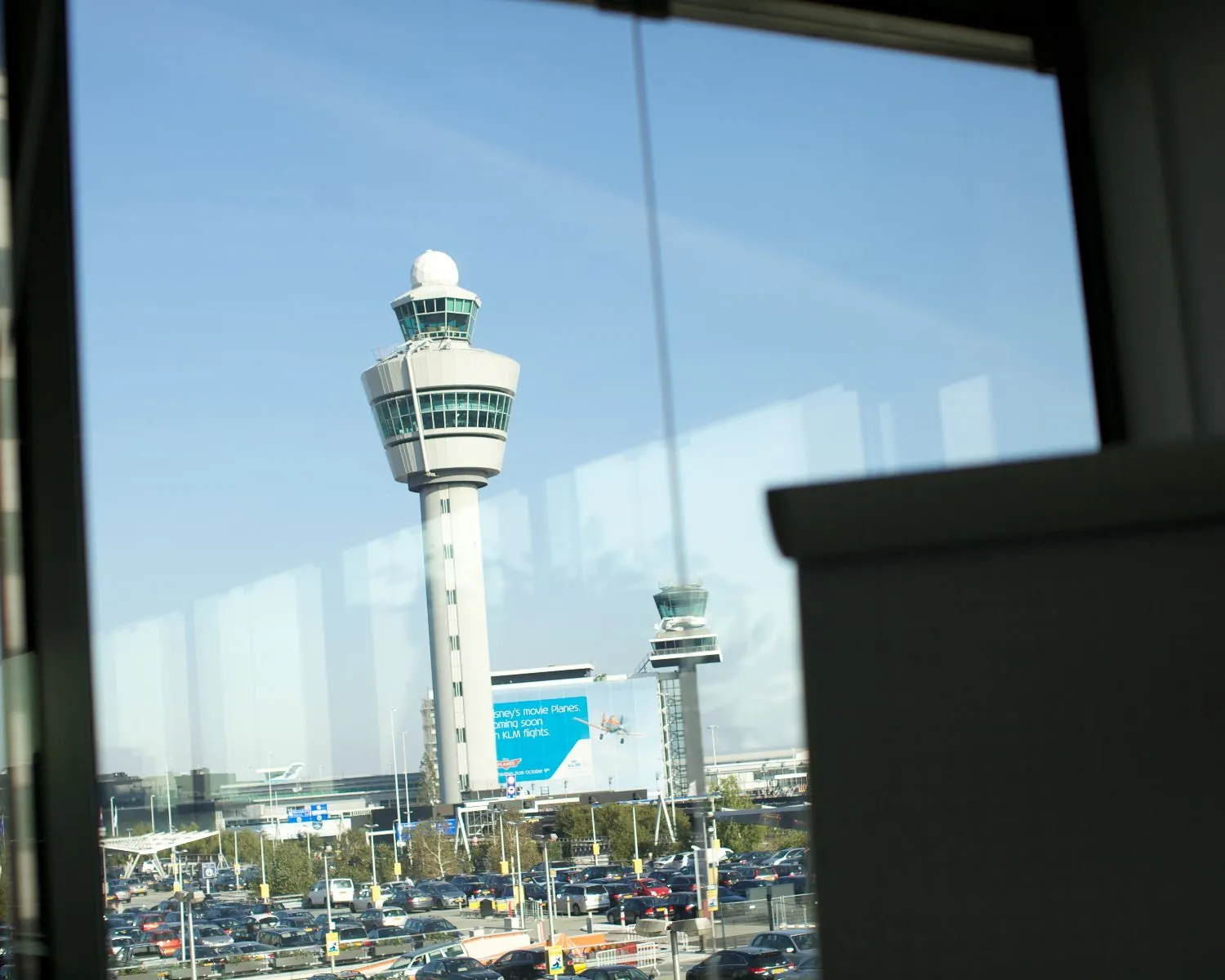 Uitzicht op de verkeerstoren van Schiphol Airport door een raam met op de voorgrond een parkeerplaats.