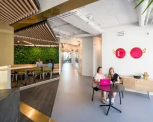 Twee vrouwen zitten aan een ronde tafel met een roze laptop in een modern kantoor met opvallende roze zonnebrilwanddecoratie.