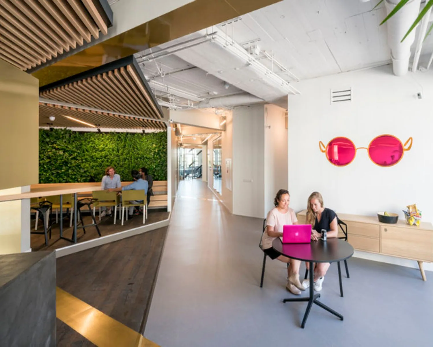Twee vrouwen zitten aan een ronde tafel met een roze laptop in een modern kantoor met opvallende roze zonnebrilwanddecoratie.