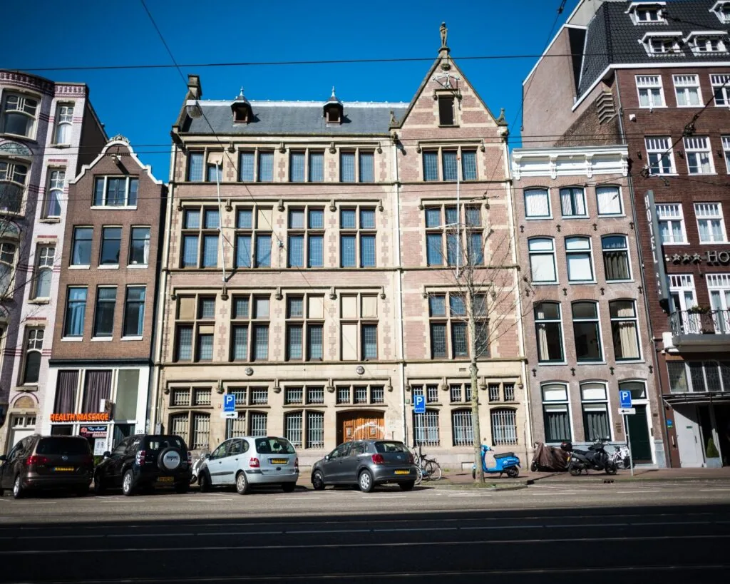 Voorgevel van een historisch pand in Amsterdam met geparkeerde auto's en fietsen langs de straat.