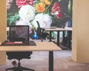 Moderne kantoorruimte met bureaus en bureaustoelen, tegen een kleurrijke bloemenmuur als achtergrond.