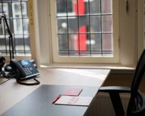 Opgeruimd bureau bij een raam met een bureautelefoon, notitieboekjes en een rode pen.