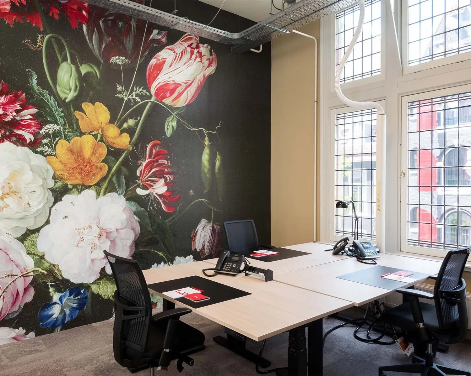 Kantoorruimte met vier bureaus en stoelen, een grote bloemenmuurschildering en ramen met glas-in-lood.