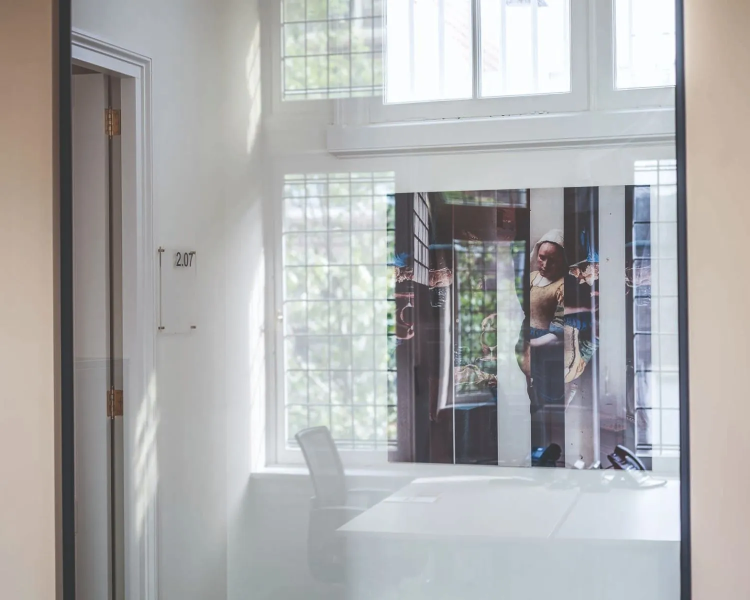 Glazen kantoorruimte met een kunstposter van Vermeers 'Melkmeisje' en kamernummer 2.07 op de muur.