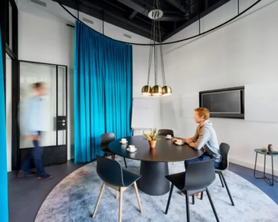 Vergaderruimte met ronde tafel, blauwe gordijnen en twee mensen, waarvan één zittend en één lopend.