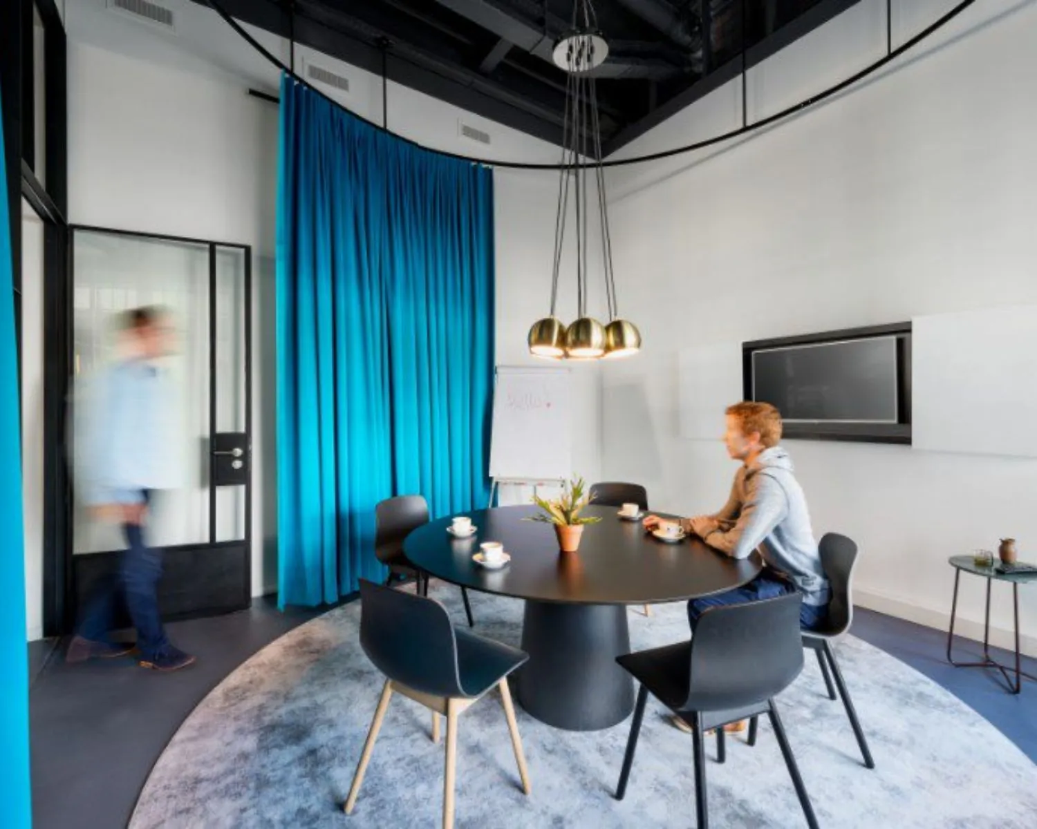 Vergaderruimte met ronde tafel, blauwe gordijnen en twee mensen, waarvan één zittend en één lopend.