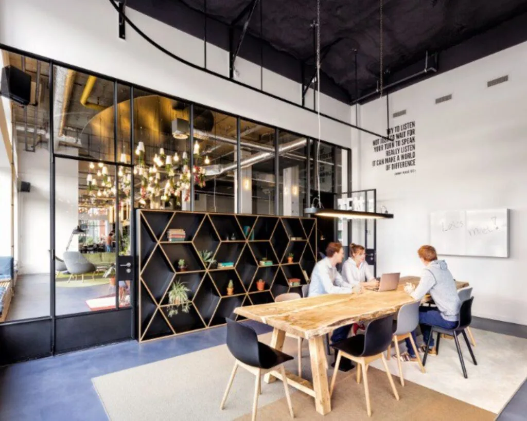 Drie mensen zitten aan een houten vergadertafel in een modern, open kantoor met glazen wanden en een opvallende hexagonale boekenkast.