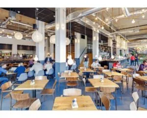 Moderne, open en drukbezochte kantine met houten tafels, industriële verlichting en mensen die eten of werken.