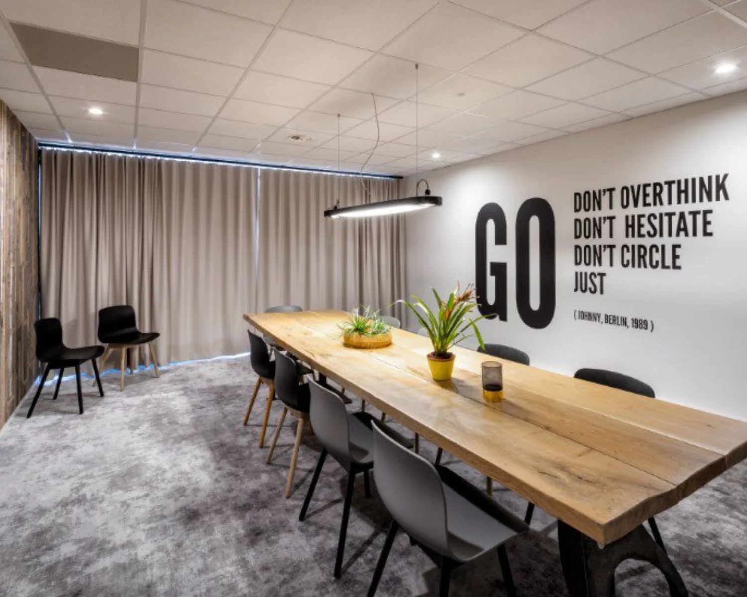 Vergaderruimte met een lange houten tafel, moderne stoelen, kamerplanten en een motiverende quote op de muur.