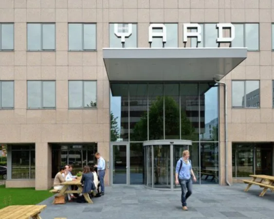 Ingang van kantoorgebouw "The Yard" met mensen aan picknicktafels en een voorbijlopende man.