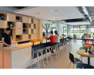 Moderne bedrijfskantine met mensen aan een bar en tafels, inclusief open keuken en grote ramen.