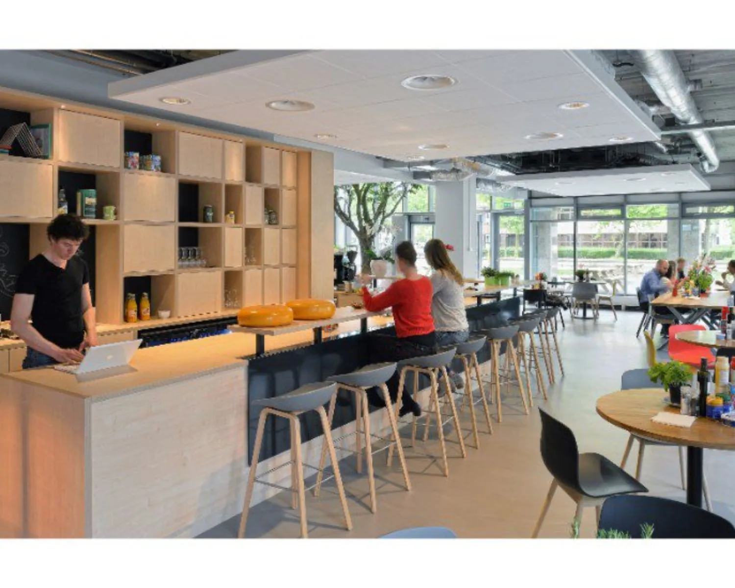 Moderne bedrijfskantine met mensen aan een bar en tafels, inclusief open keuken en grote ramen.