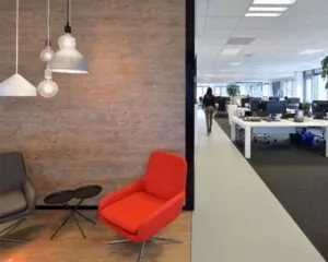 Modern kantoorinterieur met een zithoek met designstoelen en hanglampen, en een open werkruimte op de achtergrond.