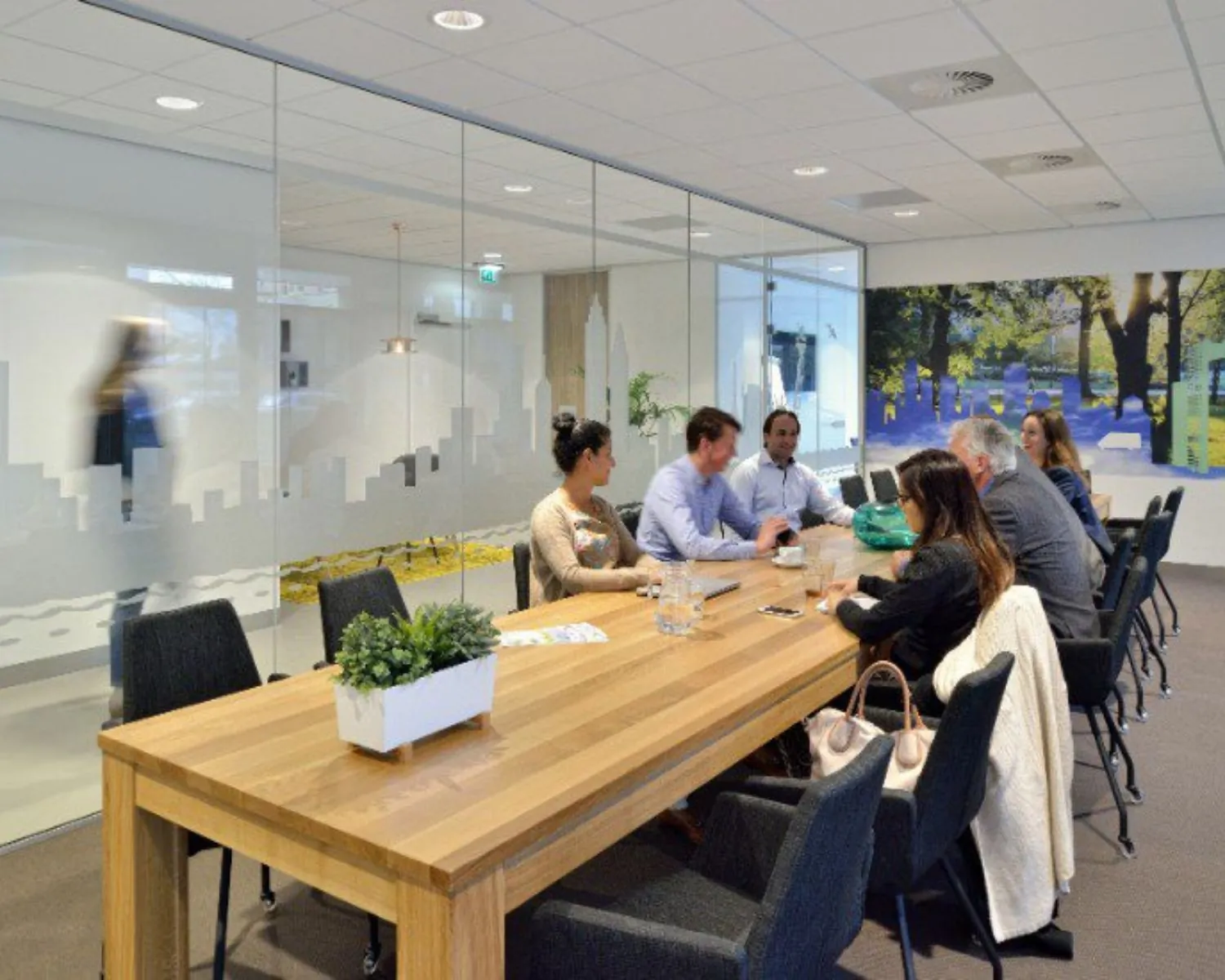 Zeven mensen zitten aan een lange houten vergadertafel in een modern kantoor met glazen wanden.