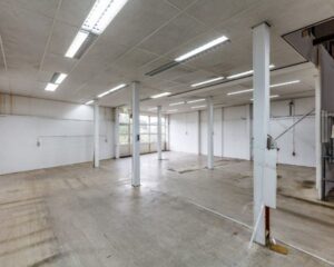 Leegstaand industrieel magazijn met betonnen vloer, witte muren en meerdere steunpilaren.