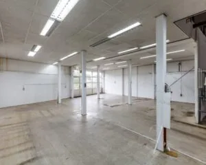 Leegstaand industrieel magazijn met betonnen vloer, witte muren en meerdere steunpilaren.