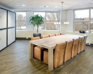 Vergaderruimte met een grote houten tafel, omringd door bruine leren stoelen en veel daglicht.