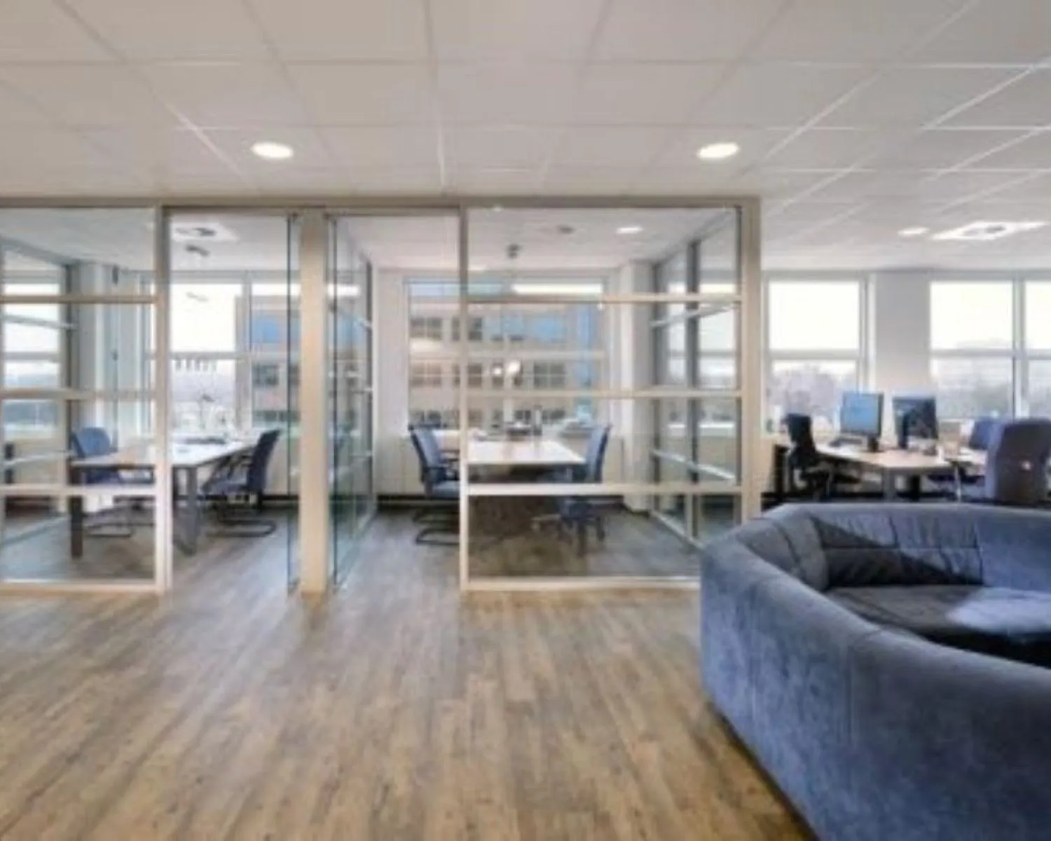 Moderne kantoorruimte met glazen vergaderruimtes, bureaus en een blauwe zithoek.