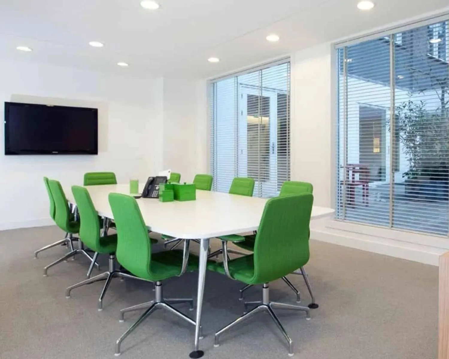 Moderne vergaderruimte met een witte tafel, groene stoelen en een flatscreen aan de muur.
