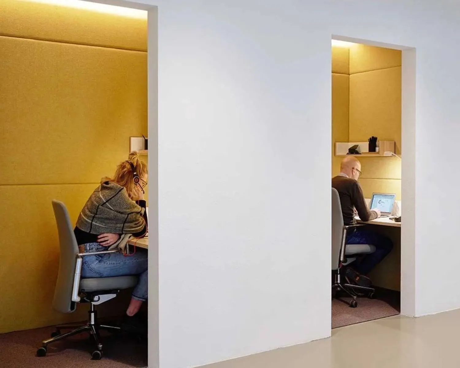 Twee mensen werken afzonderlijk in akoestisch afgezonderde werkplekken met gele wanden.
