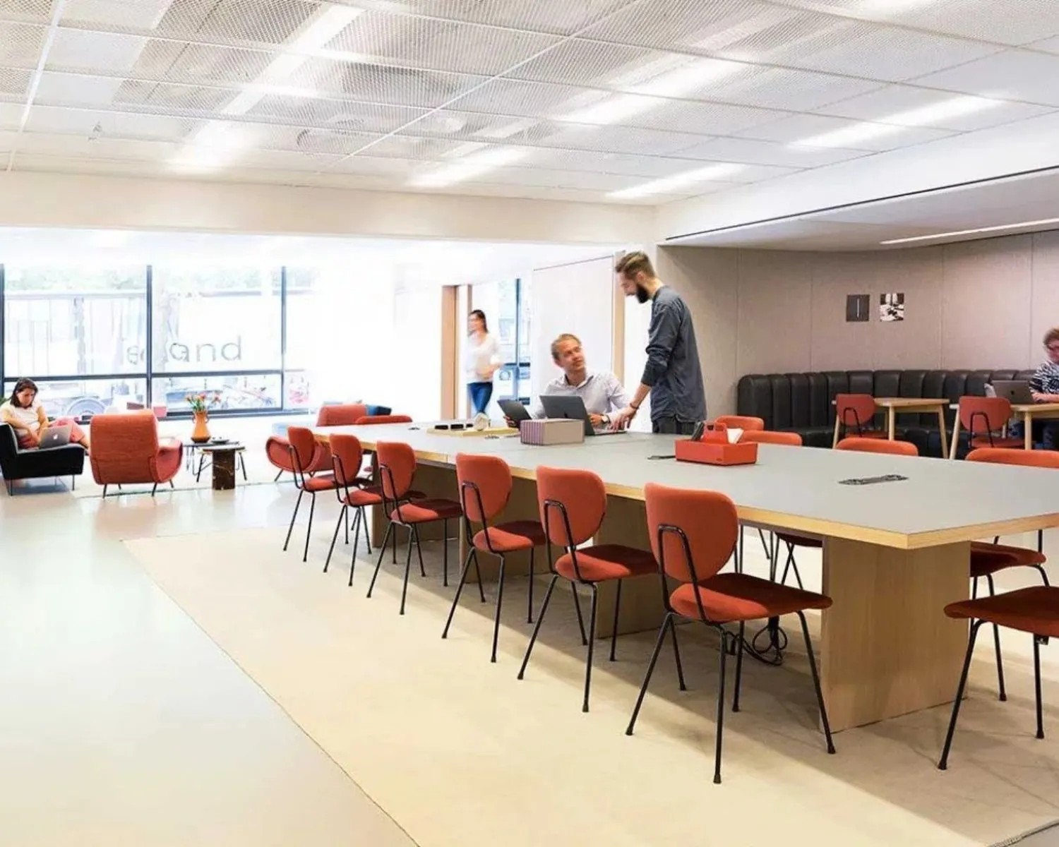 Moderne gezamenlijke werkruimte met lange vergadertafel, rode stoelen en mensen die op laptops werken.