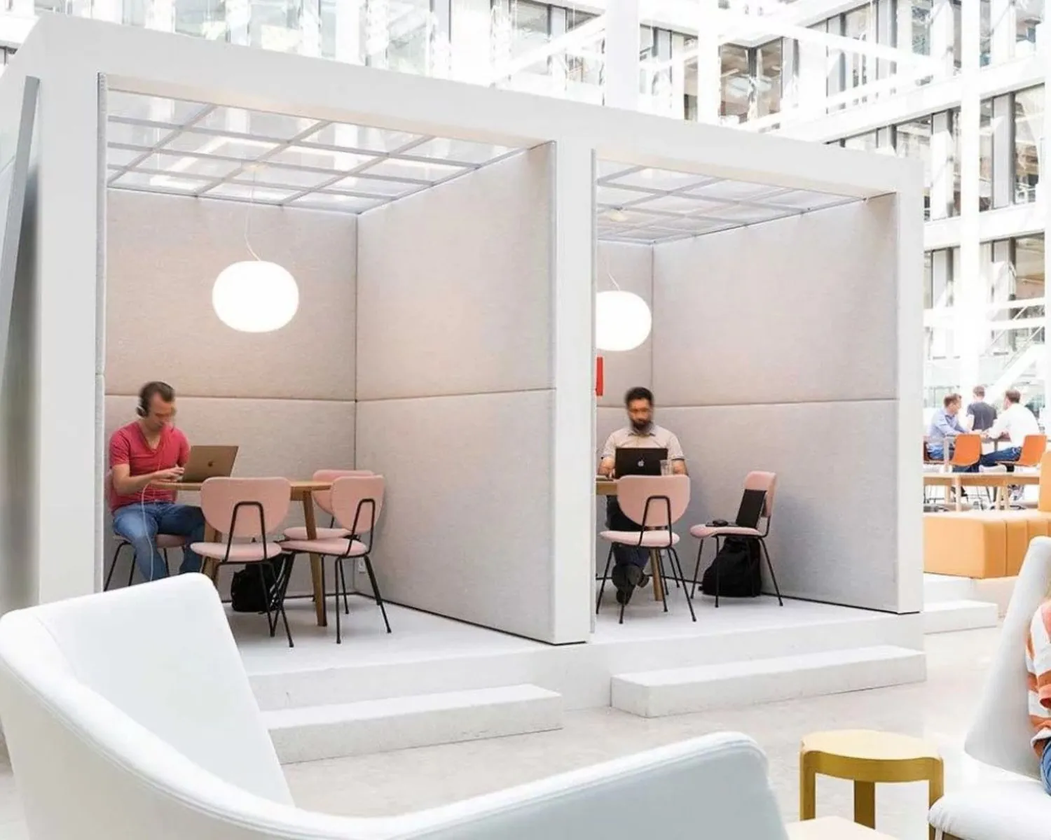 Twee mannen werken op laptops in moderne, open werkpods met zachte wanden en roze stoelen in een lichte kantooromgeving.