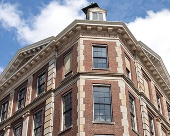 Hoekaanzicht van een klassiek bakstenen herenhuis met witte raamlijsten en sierlijke geveldetails tegen een blauwe lucht.