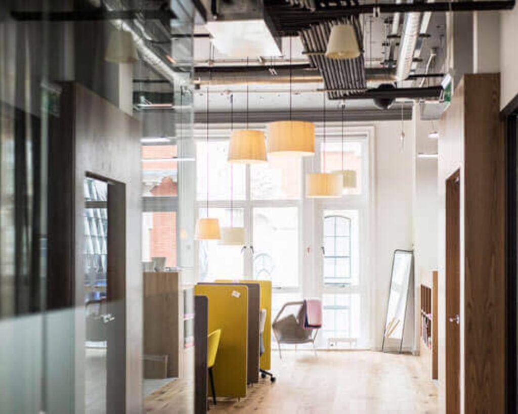 Modern kantoorinterieur met open werkplekken, gele stoelen en hanglampen, verlicht door natuurlijk daglicht.