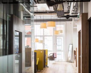 Modern kantoorinterieur met open werkplekken, gele stoelen en hanglampen, verlicht door natuurlijk daglicht.