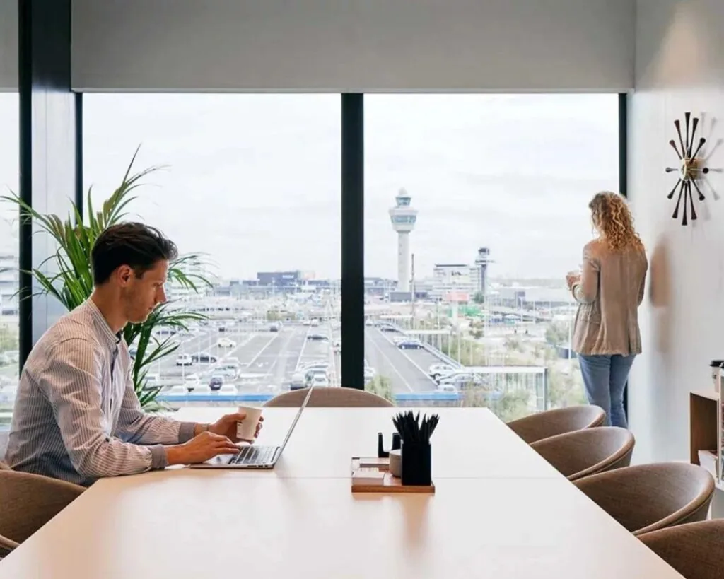 Man werkt op laptop aan vergadertafel terwijl vrouw uitkijkt op Schiphol vanachter kantoorraam.